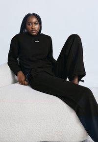 Črni pulover s kapuco z napisom "HUGO", kombiniran z ohlapnimi črnimi hlačami. Model sedi na teksturirani beli tkanini kavča, kar izžareva sproščeno udobje.