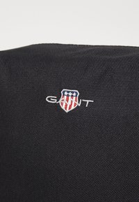 Svart stoff med en teksturert overflate, prydet med et brodert GANT-logo i hvitt og rødt, innkapslet i en skjoldform med stjerner.