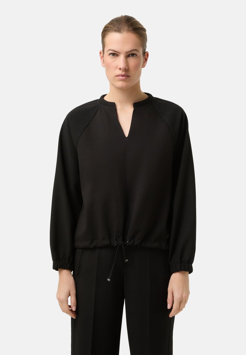 Černý pulovr s výstřihem do V, elastickými manžetami a stahovací šňůrkou v pase. Hladká látka; volný střih. V kombinaci s odpovídajícími černými kalhotami.