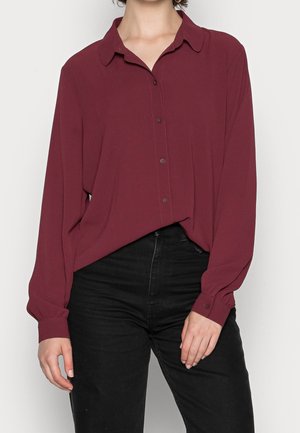 Femme portant un chemisier ample à manches longues de couleur bordeaux avec des boutons, rentré dans un pantalon noir taille haute.