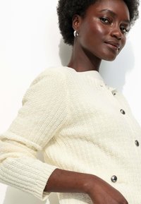 Cardigan en tricot crème avec un motif texturé, des poignets côtelés et de grands boutons sombres. Le modèle pose sur un fond blanc.