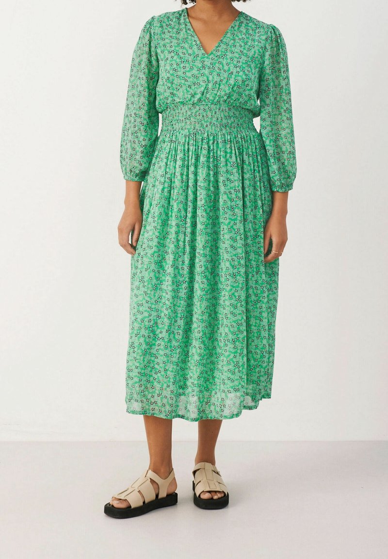 Robe midi verte à fleurs avec manches bouffantes, taille smockée et tissu léger. Associée à des sandales beiges à lanières avec semelles noires.