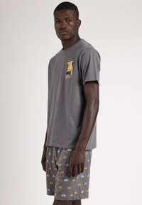 Camiseta gris con un pequeño gráfico en el pecho, combinada con pantalones cortos grises que presentan un patrón repetido de vehículos en amarillo y blanco.