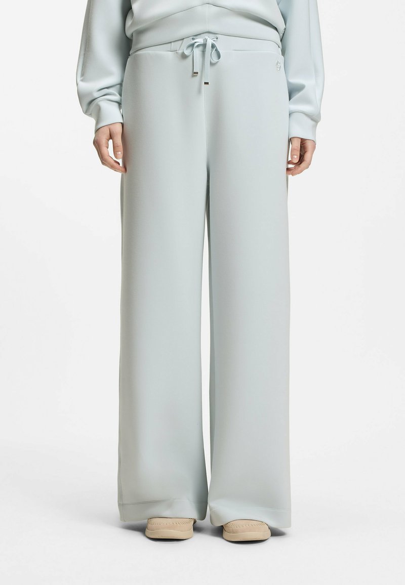 Personne portant un pantalon ample gris clair à taille élastique avec cordon de serrage et des mocassins beiges, debout devant un fond blanc.