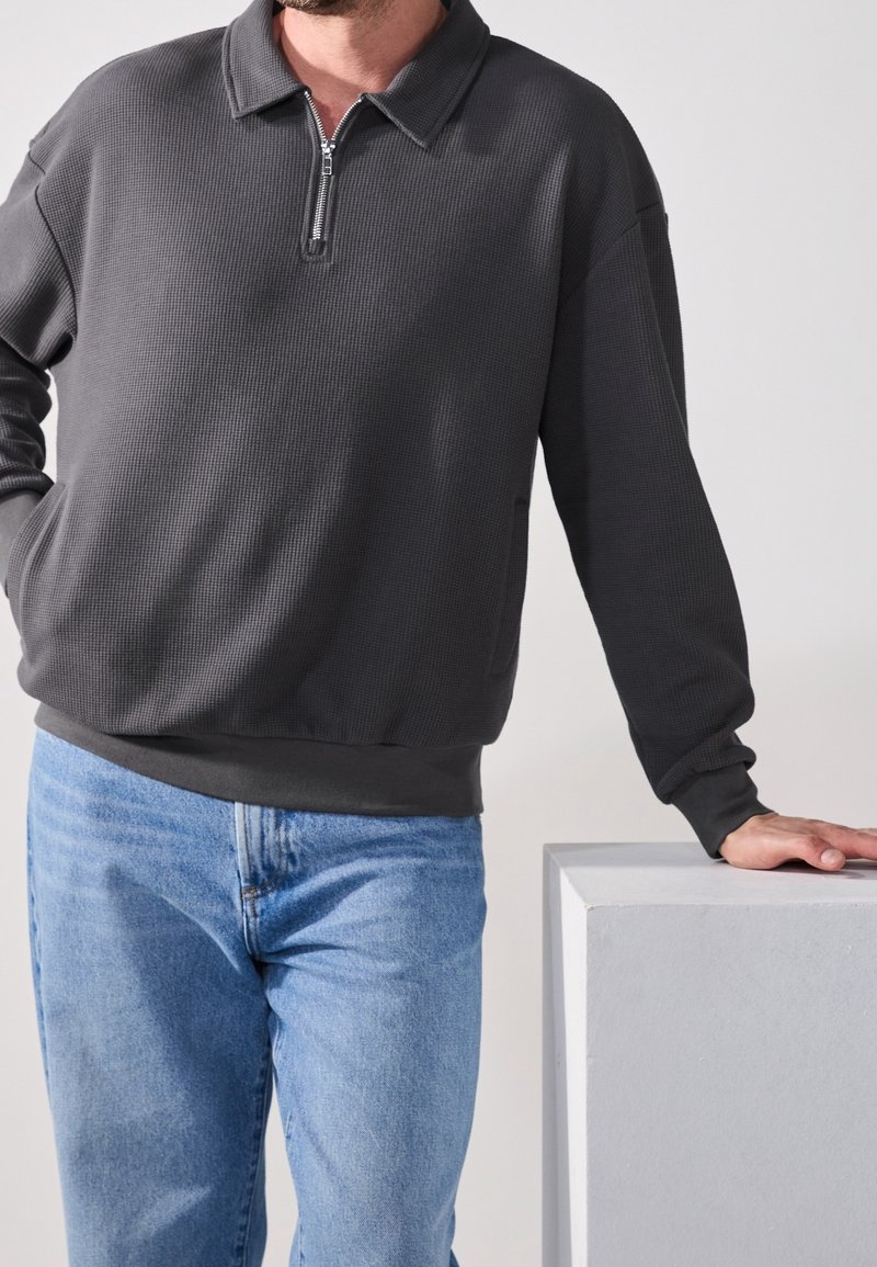 Pullover grigio con zip in tessuto strutturato, caratterizzato da un colletto e polsini a coste, abbinato a jeans di denim azzurro chiaro.