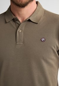 Khaki polo tričko vyrobené z texturované tkaniny, s límcem, dvouknoflíkovou légou a malým kruhovým logem s červenými a černými akcenty.