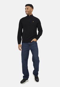 Maglione nero con zip al collo, realizzato in un tessuto liscio, abbinato a jeans blu a gamba dritta e scarpe da ginnastica nere. Dettaglio del logo dorato sul petto.