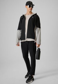 Homme en tenue de sport noire et grise, casquette et lunettes de soleil réfléchissantes, debout, tenant une gourde noire et regardant sur le côté.