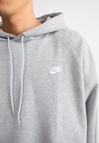 Personne portant un sweat à capuche Nike gris clair avec cordons, montrant le haut du torse et le logo de la marque sur la poitrine.