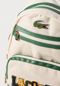Bežinė audinė kuprinė su žaliais odiniais akcentais, su krokodilo logotipu ir daugiaspalviu "LACOSTE" tekstu tekstūriniu siuvinėjimu.