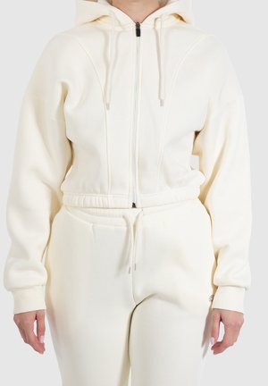 Osoba ubrana w kremową bluzę z kapturem na zamek błyskawiczny z troczkami oraz dopasowane, wysokowstawe spodnie dresowe z troczkiem w pasie.