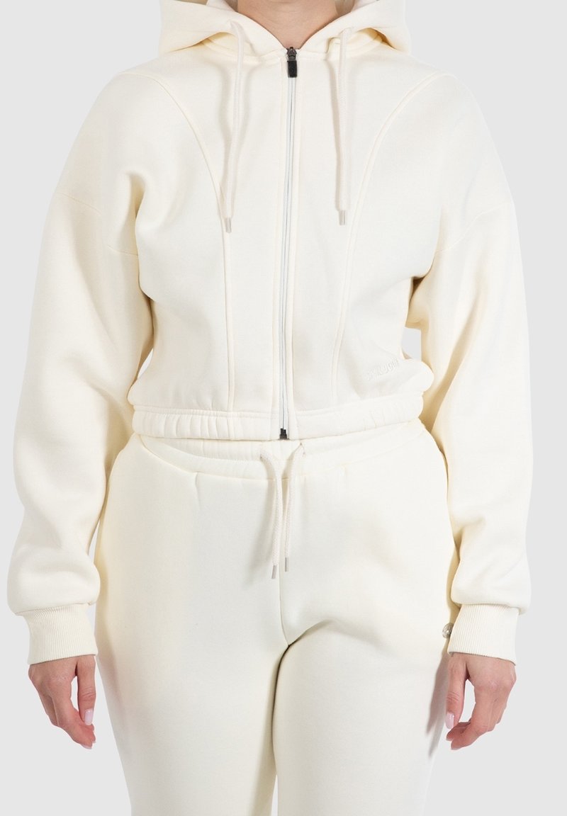 Personne portant un sweat à capuche crème zippé avec cordons et un pantalon de jogging taille haute assorti avec cordon à la taille.