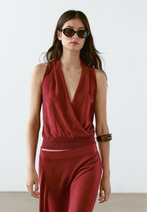 Red knit wrap top with deep V-neckline and ribbed hem. Paired with matching wide-leg pants. Worn with black sunglasses and a bangle bracelet.