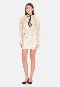 Blusa a motivo crema con dettagli neri e fiocco, abbinata a una gonna crema coordinata. Modella indossa scarpe con tacco nere.