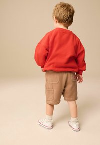 Toddler wearing a red sweatshirt, beige shorts with pockets, cream socks, and white sneakers, standing with their back facing forward.