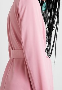 Personne portant un manteau rose ceinturé, vue de profil, avec de longs cheveux tressés et des boucles d'oreilles géométriques bleu sarcelle, visibles sur un fond uni.