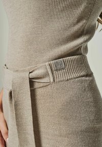 Beiges Strickkleid mit gerippter Textur, einem Bindegürtel und einem Markenpatch an der Taille. Der Stoff wirkt weich und figurbetont.