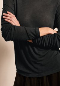 Schwarzes Langarmshirt aus einem weichen, leichtem Stoff mit rundem Ausschnitt und leicht gerafftem Saum, getragen mit verschränkten Armen.