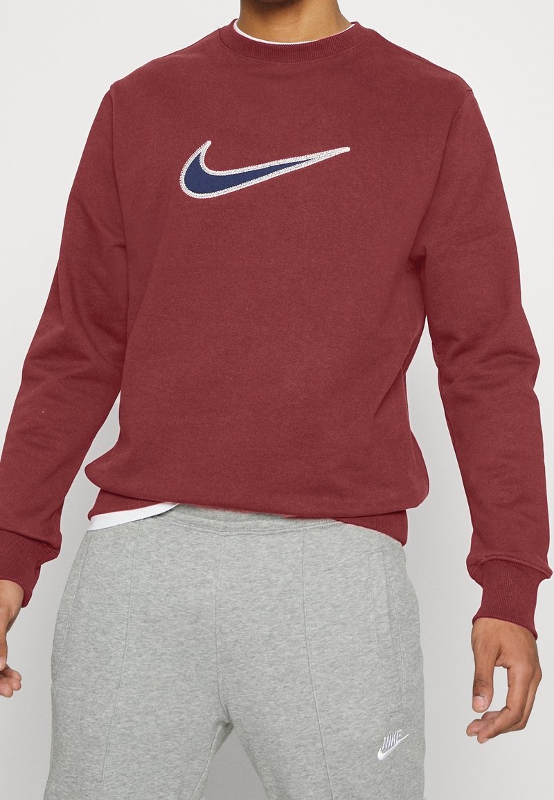 Sudadera de cuello redondo en color burdeos, hecha de una mezcla de algodón, con un logo bordado de Nike en blanco y azul marino en el pecho. Pantalones de chándal grises visibles por debajo.