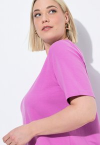 Maglietta a maniche corte, colletto rotondo, in cotone rosa brillante. Tessuto liscio, forma aderente, dettagli cuciti visibili su maniche e orlo.