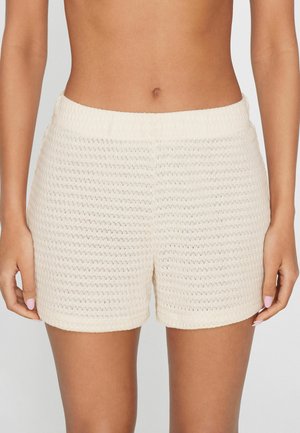 Kobieta w kremowych plecionych szortach z elastycznym pasem, stojąca z ramionami swobodnie opuszczonymi wzdłuż ciała na jednolitym tle.