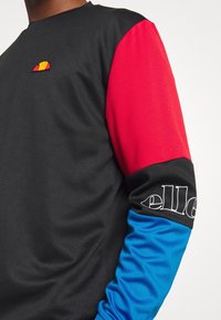 Svart sweatshirt med röd övre ärm, blå nedre ärm, liten färgglad logotyp på bröstet och vit "ellesse"-text på svart mid-ärmsband.