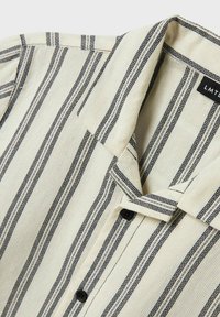 Teksturert kremfarget skjorte med svarte horisontale striper, spiss krage og to svarte knapper foran. Merkeetikett synlig ved halsen.