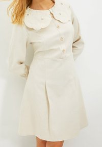 Vestido beige con un cuello de encaje con borde festoneado, cierre delantero con botones, mangas largas y una falda acampanada con pliegues sutiles. Textura de tela suave.