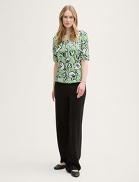 Grüne Bluse mit kurzen Ärmeln und schwarzem Blumenmuster, kombiniert mit schwarzen weit geschnittenen Hosen und schwarzen Ballerinas. Einfarbiger Hintergrund.