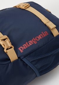 Tamsiai mėlynas kuprinės modelis su smėlio spalvos diržais, tinklelio kišenėmis ir raudonu "Patagonia" logotipu. Pagaminta iš patvarios medžiagos, su reguliuojamais užsegimais saugiam uždarymui.