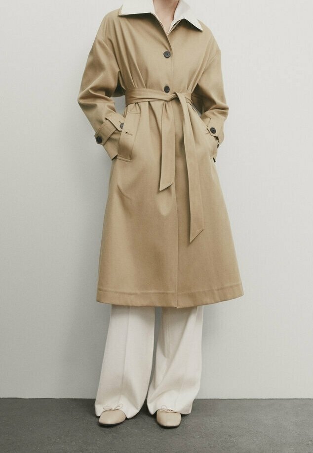 Personne portant un trench-coat beige ceinturé avec des boutons noirs, un pantalon blanc à jambes larges et des ballerines beiges, debout devant un fond uni.