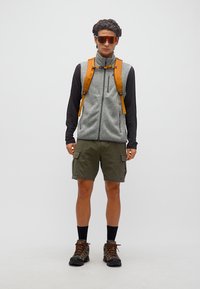 Νέος άνδρας με πορτοκαλί γυαλιά ηλίου, γκρι γιλέκο από φλις, μαύρο μακρυμάνικο πουκάμισο, χακί βερμούδα με τσέπες, ορειβατικά παπούτσια και ορατές πορτοκαλί ράντες σακιδίου.