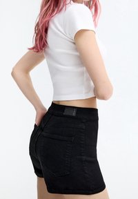 Mujer con cabello rosa que lleva una camiseta blanca de corte cropped y pantalones cortos de mezclilla negros, de pie con las manos en los bolsillos contra un fondo blanco.