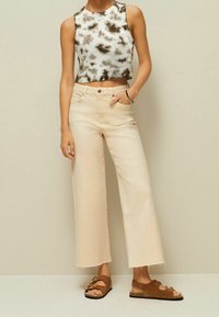 Donna che indossa un top corto senza maniche nero e bianco a pois, jeans a vita alta beige con gamba larga e sandali marroni con fibbia su sfondo neutro.