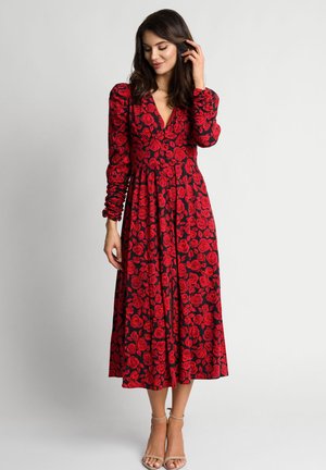 Kobieta w czarnej, midi sukience z długimi rękawami i czerwonymi wzorami róż, stojąca z jedną ręką dotykającą włosów, w beżowych szpilkach.