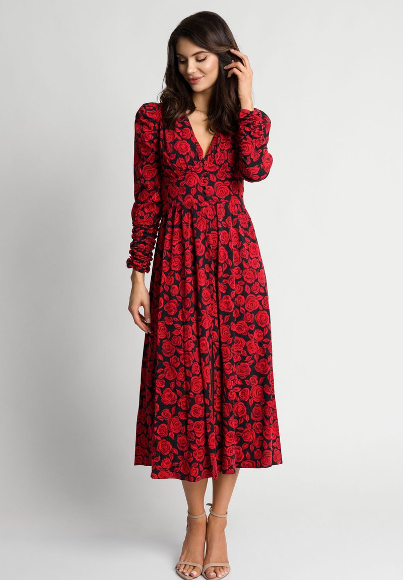Kobieta w czarnej, midi sukience z długimi rękawami i czerwonymi wzorami róż, stojąca z jedną ręką dotykającą włosów, w beżowych szpilkach.