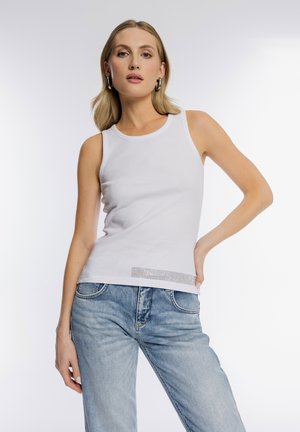 Blondynka w srebrnych kołach, białym bezrękawnym topie i jasnoniebieskich dżinsach, pozująca z jedną ręką na biodrze na białym tle.