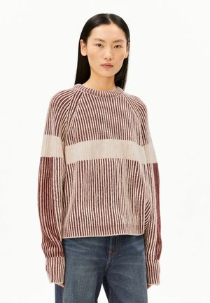 Femme aux cheveux noirs lisses portant un pull rayé à manches longues beige et bordeaux avec un jean bleu, debout sur un fond blanc.