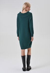 Abito lavorato a maglia teal con un ampio colletto ripiegato, maniche lunghe e polsini a costine; abbinato a stivali neri in pelle alti con dettagli dorati.