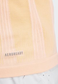 Camiseta deportiva Aeroready en un tono melocotón claro, con tejido texturizado, patrón de rayas verticales y dobladillo acanalado. Puntos amarillos de acento cerca del área del cuello.