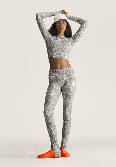 Mujer con top corto y leggings con estampado de serpiente en gris y blanco, gorra blanca y zapatos naranja brillante, de pie con los brazos levantados detrás de la cabeza.