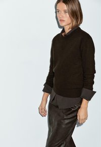 Maglione di lana marrone con maniche lunghe e scollo tonda, indossato sopra una camicia marrone scuro. Abbinato a una gonna di pelle marrone.