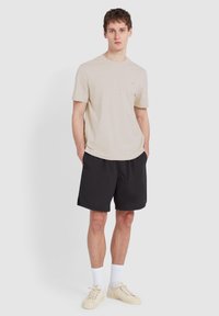 Beige T-Shirt mit kurzen Ärmeln, schwarze Shorts, weiße Knöchelsocken und cremefarbene Sneaker. Das Model steht mit den Händen in den Taschen vor einem schlichten Hintergrund.