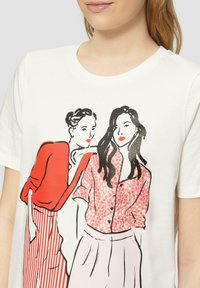 T-shirt blanc présentant un dessin en ligne noir de deux femmes vêtues de tenues rouges et roses, avec des motifs floraux et rayés. Manches courtes, col rond.