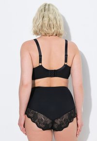 Zwart bh en hooggeknoopte slipset, met een gladde stof en kantafwerking. De slip heeft een golvende rand en een geometrisch kantpatroon.