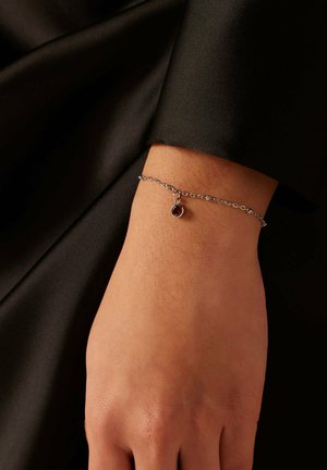 Silver chain bracelet featuring a round red gemstone pendant, worn on a bare wrist, against a dark background. Soft texture visible.