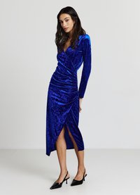 Abito in velluto blu con maniche lunghe, design avvolgente, arricciature laterali e spacco alto. Scarpe con tacchi neri e punta affilata completano il look.