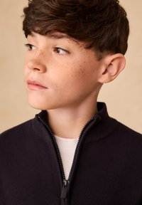 Tumesinine lukuga kampsun kõrge kraega, soonilise tekstuuriga ja musta lukuga. Kanditud valge T-särgi peal. Näha on täpilist nahka.