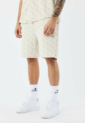Pantalones cortos beige con un diseño de diamantes, hechos de una tela texturizada. Combinados con zapatillas blancas y calcetines con un logo negro.