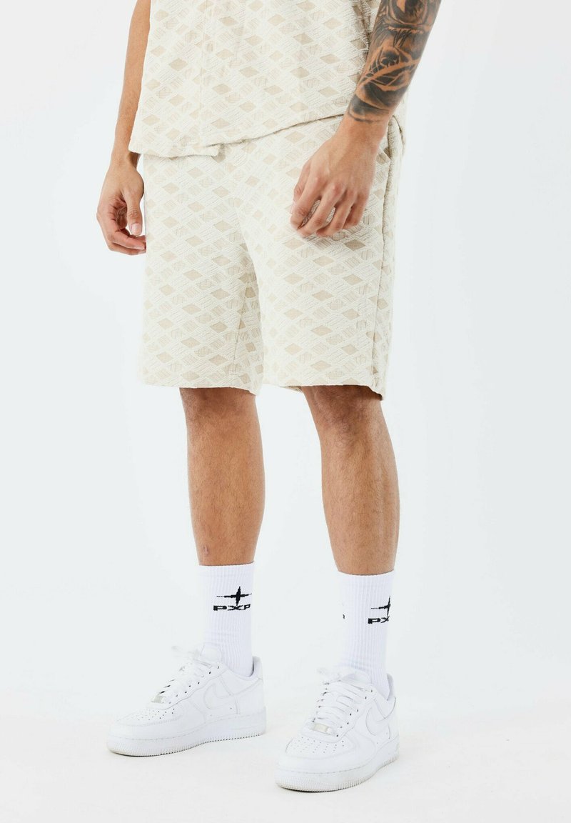 Shorts beiges à motif avec un design en losange, en tissu texturé. Associés à des sneakers blanches et des chaussettes avec un logo noir.